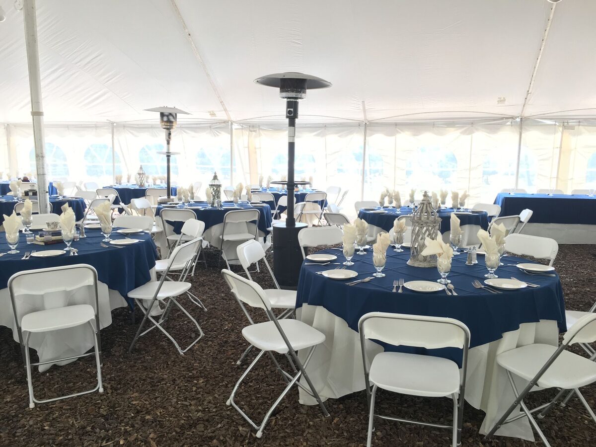 Reception seating inside a pole tent with round tables, white folding chairs, navy linens, china, glassware, and outdoor heaters